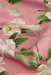 Tkanina s cvetličnim vzorcem, ki prikazuje bele cvetove in zelene liste na roza ozadju. Vsebuje blagovno znamko v zeleni barvi: "by Ted Baker."