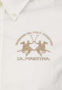 Bílá košile s límečkem, zdobená béžovým vyšitým logem polo s dvěma jezdeckými postavami a textem, na hladké bavlněné textuře.