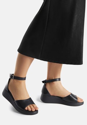 Czarne skórzane sandały na koturnie mają otwarty przód, pasek wokół kostki z klamrą oraz teksturowaną podeszwę zapewniającą przyczepność i komfort.