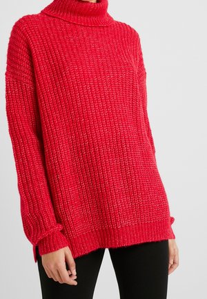 Czerwony, długi sweter z dzianiny z golfem, noszony z czarnymi spodniami, widoczny od ramion do bioder na jednolitym tle.