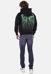 Felpa nera in cotone con un vivace design grafico verde sul retro, abbinata a jeans blu e sneakers bianche.