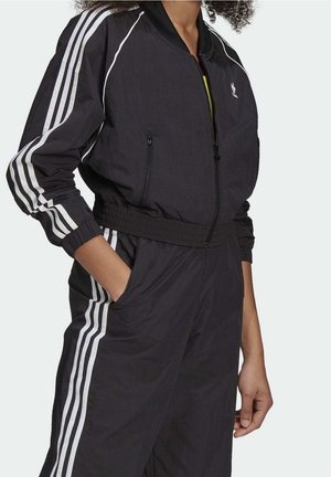 Kvinde iført sort Adidas træningsdragt med hvide tre-stribe detaljer på ærmer og bukseben, hænderne i lommerne, stående mod en ensfarvet baggrund.