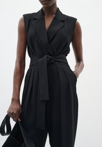 Combinaison noire à ceinture avec un design enveloppant, sans manches et poches latérales. Tissu lisse avec un éclat subtil et une silhouette structurée.