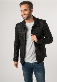 Giubbotto di pelle nera con una texture liscia, dotato di cerniere, bottoni a scatto e cuciture a contrasto. Indossato sopra una camicia bianca e jeans blu.