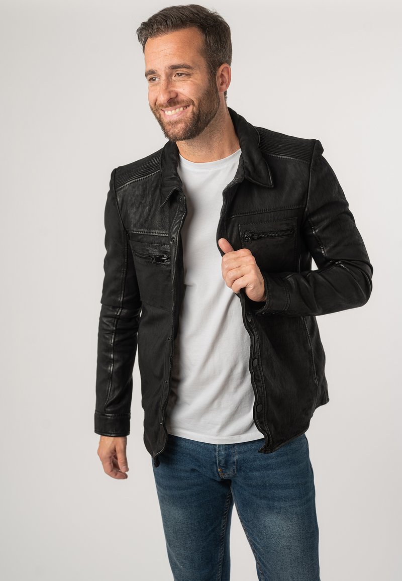 Giubbotto di pelle nera con una texture liscia, dotato di cerniere, bottoni a scatto e cuciture a contrasto. Indossato sopra una camicia bianca e jeans blu.
