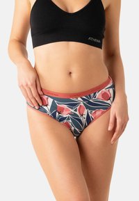 Femme portant un soutien-gorge de sport noir et un sous-vêtement de bikini à motifs floraux avec des bords corail sur un fond uni.
