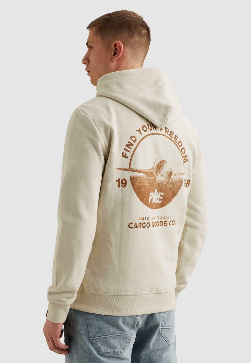 Homme portant un sweat à capuche beige avec la capuche relevée, un motif d'avion et le texte "Trouvez votre liberté" au dos, associé à un jean bleu clair.