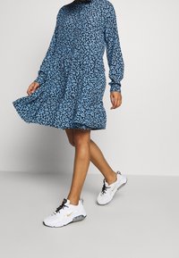 Blaues Blumenmuster-Kleid mit durchgehender Knopfleiste, langen Ärmeln und tailliertem Oberteil, kombiniert mit weißen Turnschuhen mit schwarzen und goldenen Akzenten.