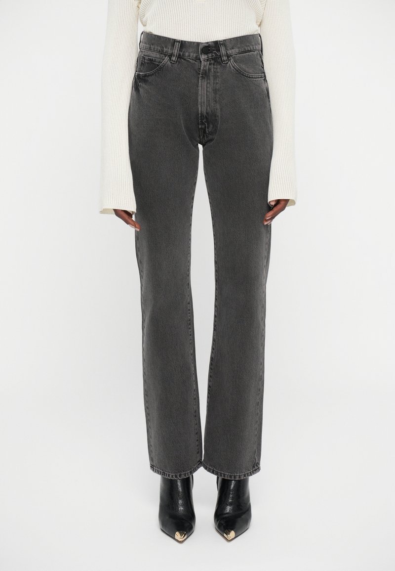 Persona que lleva pantalones vaqueros negros de cintura alta y rectos, un suéter crema de manga larga con canalé y botas negras de punta afilada con detalles dorados.