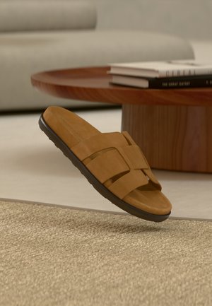 Brun ruskind slidssandal med sort sål svævende over beige tekstureret tæppe, rundt træ sofabord og sofa i baggrunden.