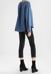Femme portant une chemise en denim bleue ample, un jean noir court avec des ourlets effilochés, et des bottines noires à talons, debout devant un fond blanc.