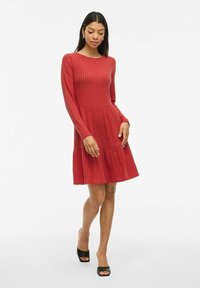 Rotes geripptes Kleid mit langen Ärmeln, rundem Ausschnitt, tailliertem Oberteil und schwingendem Rock. Kombiniert mit schwarzen offenen Schuhen.
