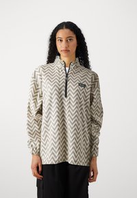 Quarter-zip pullover in light beige with a zigzag pattern in darker beige, featuring elastic cuffs and a logo patch on the chest.