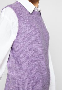 Helllila gestrickte Weste mit strukturiertem Muster, runder Ausschnitt und ärmellosem Design, über einem weißen Hemd getragen.
