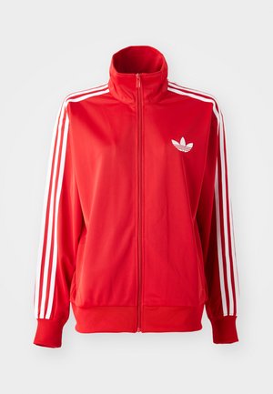 Sarkans zip-up sporta žakets, izgatavots no gludas auduma, ar baltām trim svītrām uz piedurknēm un mazu baltu logo uz krūtīm.