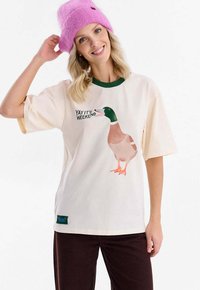 Krémově bílé bavlněné tričko se zeleným límcem, zdobené grafikou ptáka a textem „YAY IT'S WEEKEND.“ Krátké rukávy, v kombinaci s hnědými kalhotami.