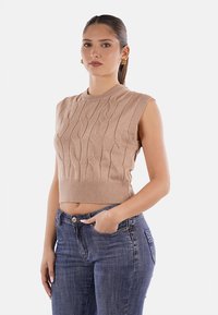 Top in maglia beige senza maniche con un motivo a treccia testurizzato, design cropped e orlo a coste, abbinato a jeans blu scuro e un atteggiamento casual.