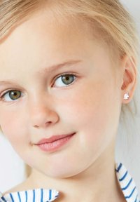 Kleine zilveren eenhoornoorbellen met witte emaille details, gecombineerd met een blauw-wit gestreept shirt. Korte blonde haren omlijsten het gezicht.