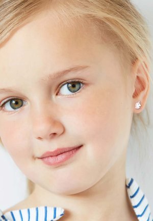 Kleine zilveren eenhoornoorbellen met witte emaille details, gecombineerd met een blauw-wit gestreept shirt. Korte blonde haren omlijsten het gezicht.