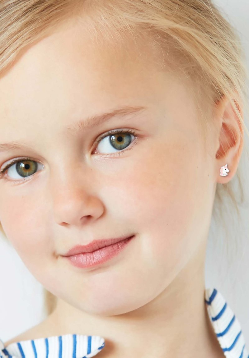 Kleine zilveren eenhoornoorbellen met witte emaille details, gecombineerd met een blauw-wit gestreept shirt. Korte blonde haren omlijsten het gezicht.