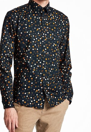 Man draagt een donker overhemd met gekleurde polkadots en beige broek, handen ontspannen langs het lichaam.