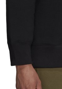 Svart sweatshirtmudd med ribbad textur och figurnära design. Den synliga handen visar detaljerad hudtextur mot den olivgröna tyget nedan.