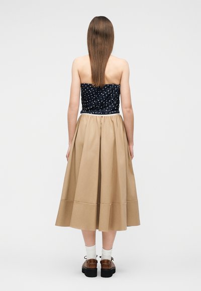 Γυναίκα που στέκεται με την πλάτη γυρισμένη, φοράει μπλε σκούρο τοπ χωρίς τιράντες με πουά, μπεζ midi φούστα, λευκές κάλτσες και καφέ παπούτσια σε απλό φόντο.