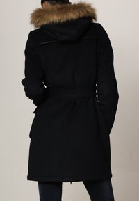 Manteau noir avec une capuche doublée en fourrure, taille ceinturée et manches longues. Présente une texture lisse et une coupe jusqu'au genou. Détails de couture subtils.