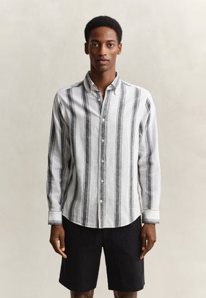 Young man standing wearing a white button-up shirt with vertical black stripes and black shorts against a plain background.