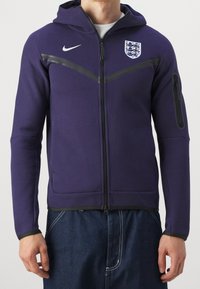 Sweat à capuche zippé violet en matériau doux, avec un motif à rayures noires, des poches avant et une poche latérale zippée. Logo Nike et écusson de l'Angleterre.