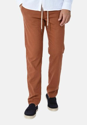 Uomo che indossa pantaloni con coulisse color ruggine, camicia bianca e scarpe nere senza lacci, in piedi davanti a uno sfondo semplice.