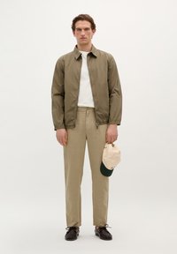 Oliven grøn letvægtsjakke, hvid t-shirt, beige linnedbukser, holder en lys beige kasket med grøn skygge, og mørke sko.