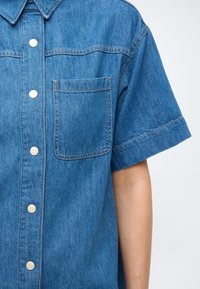 Modrá džínsová košeľa s krátkym rukávom má zapínanie na gombíky vpredu, náprsné vrecko a decentné švíkové detaily. Mäkká textúra a pohodlný strih.