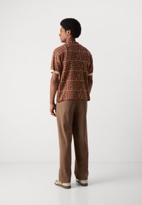 Short-sleeve brown shirt with large "HEATH" text pattern, cream trim at sleeves, paired with brown trousers and light footwear.