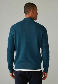 Türofanfarbener Strickpullover mit hohem Kragen, strukturiertem Stoff, gerippten Bündchen und Saum, über einem weißen Hemd getragen. Sichtbare Nähte an den Schultern.