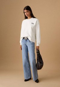 Hanorac alb cu imprimeu "ECHOES FREEDOM", blugi de culoare albastru deschis cu croială largă, cizme negre cu toc și ghete la gleznă, și o geantă neagră strânsă.