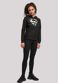 Black hoodie with a large silver logo on the chest, complemented by black leggings and black athletic shoes. Smooth material, casual fit.