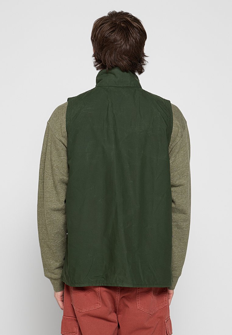 Person, der eine grüne ärmellose Jacke über einem langärmeligen olivgrünen Hemd und roten Cargo-Hosen trägt, steht mit dem Rücken zu einer einfarbigen Hintergrund.