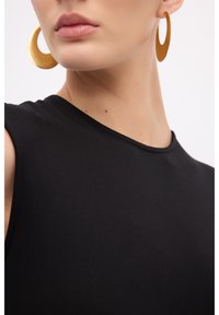 Top sans manches noir avec un col rond, agrémenté de grandes boucles d'oreilles en or, mettant en avant des textures lisses et un design simple.