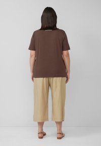 Bruine katoenen t-shirt met korte mouwen heeft een subtiele tekstprint op de achterkant. Gecombineerd met beige cropped broeken en bronzen sandalen.