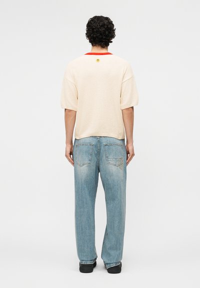 Homme aux cheveux bouclés portant un jean bleu clair ample et un pull à manches courtes crème avec un bord rouge, debout de dos sur un fond blanc.