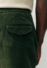 Dark green corduroy pants with a back pocket featuring a flap and button closure. Shallow ribbed texture and fitted waist.