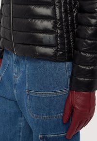 Černá péřová bunda s lesklým povrchem, s vodorovným prošíváním a malým logem. Červené kožené rukavice na pozadí z denimu.