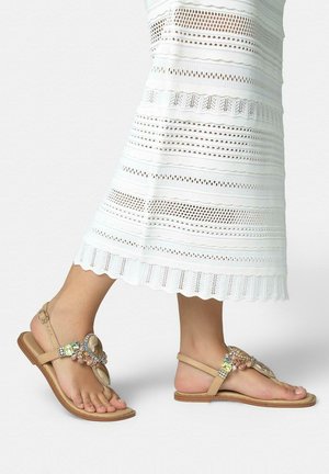 Voeten met beige T-band sandalen met kralen, gecombineerd met een witte midi-rok met een kanten, geschulpte zoom en patroonvormige uitsparingen.