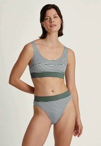 Woman wearing a black and white striped bikini with green bands, standing against a plain light background, looking slightly towards the camera.