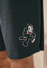 Svarta bomullsshorts med en vit tecknad kattgrafik i en löpande position, med ett vänligt uttryck och röda detaljer på munnen.