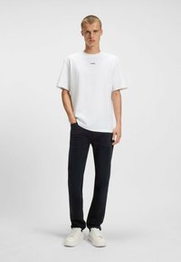T-shirt bianca a maniche corte con un logo nero sul petto, abbinata a pantaloni scuri e sneakers bianche. Design semplice e pulito.