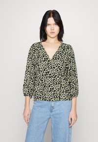 Zelena bluza sa leopard printom u crnoj boji, s dubokim V-izrezom, dugim napuhanim rukavima i skupljenim strukom, uparena s plavim farmerkama.