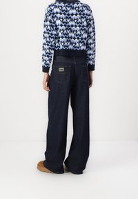 Henkilö, joka pukeutuu tummiin leveisiin farkkuihin, joissa on takataskut, ja siniseen kuviollesvettiin, seisoo yksinkertaisella valkoisella taustalla.