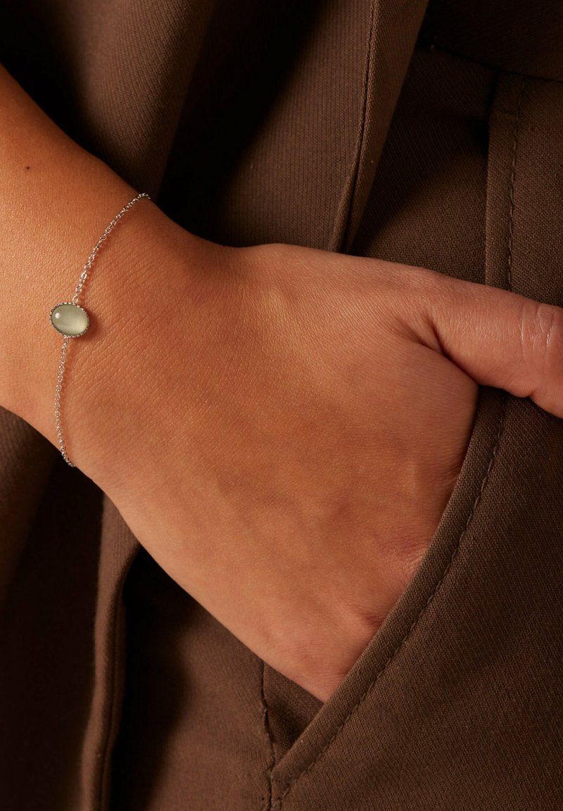 Zilveren kettingarmband met een ovale, lichtgroene steen, gedragen op een hand die in een bruine zak rust met zichtbare stoftextuur.